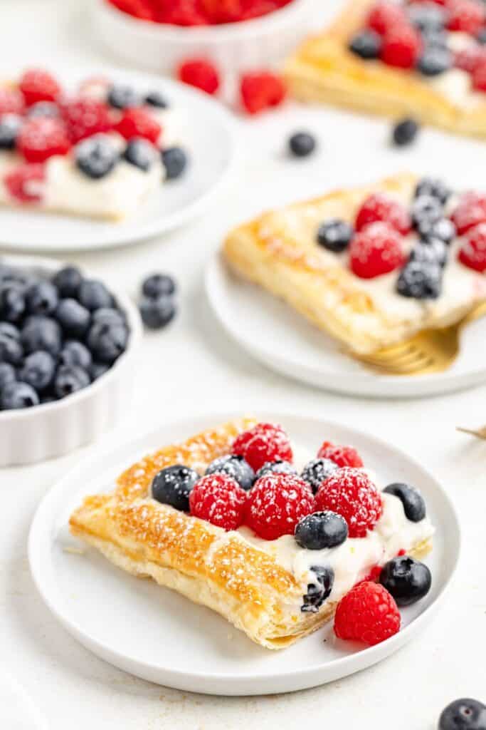 Square puff pastry pastry with cream cheese filling, raspberries, and blueberries on a white plate; extra plates and berries behind.