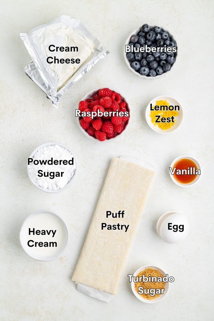 Top-down view of labeled berry tart ingredients: cream cheese, berries, lemon zest, sugars, heavy cream, puff pastry.