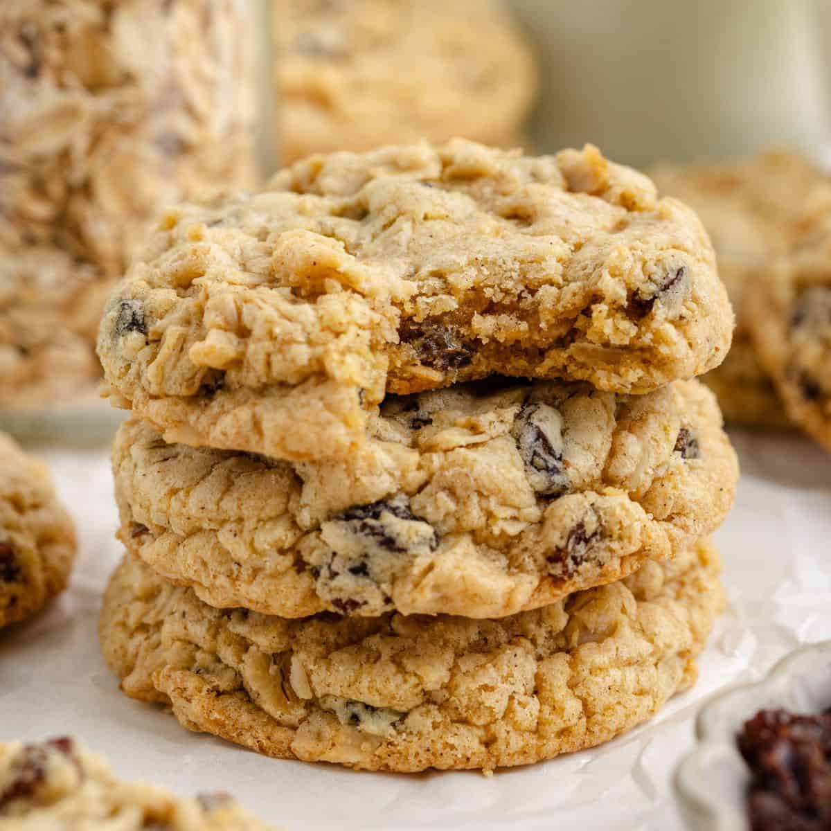 A stack of three oatmeal raisin cookies on parchment paper, with oats and additional cookies behind.