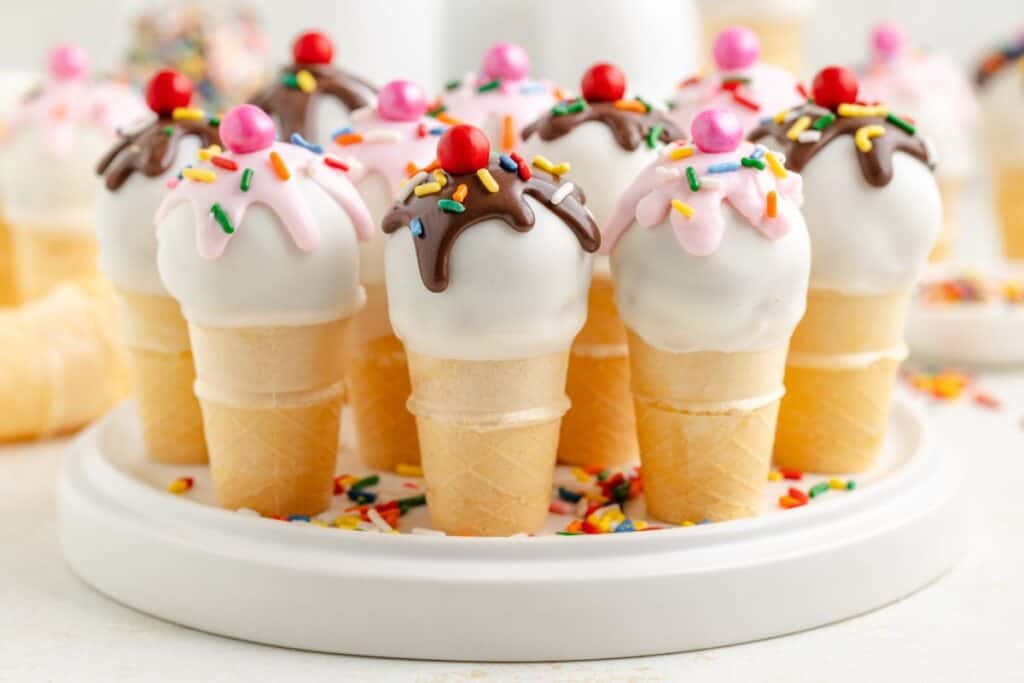 Cake pop ice cream cone treats with colorful coatings, sprinkles, and red candy on a white plate.