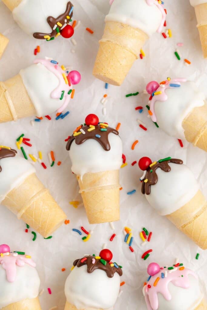 Mini ice cream cone cake pops with white chocolate coating, sprinkles, chocolate drizzle, and red candy tops on white surface.