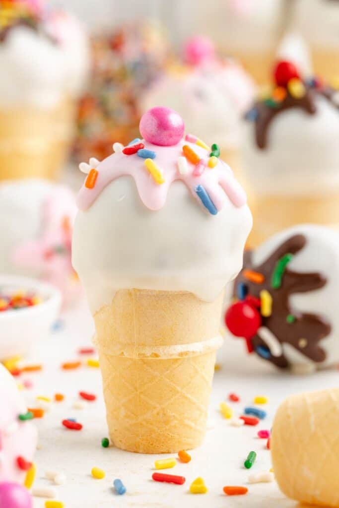 Ice cream cake pop with pink, white icing, sprinkles, and round candy, surrounded by similar decorated treats.