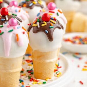 Mini ice cream cone cake pops with white coating, chocolate drizzle, sprinkles, and candy toppings arranged on a plate.