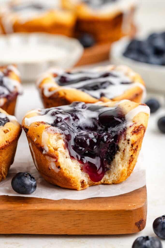 Mini blueberry pie with cinnamon roll crust, missing a bite, on wooden board; whole blueberries and pastries in background.