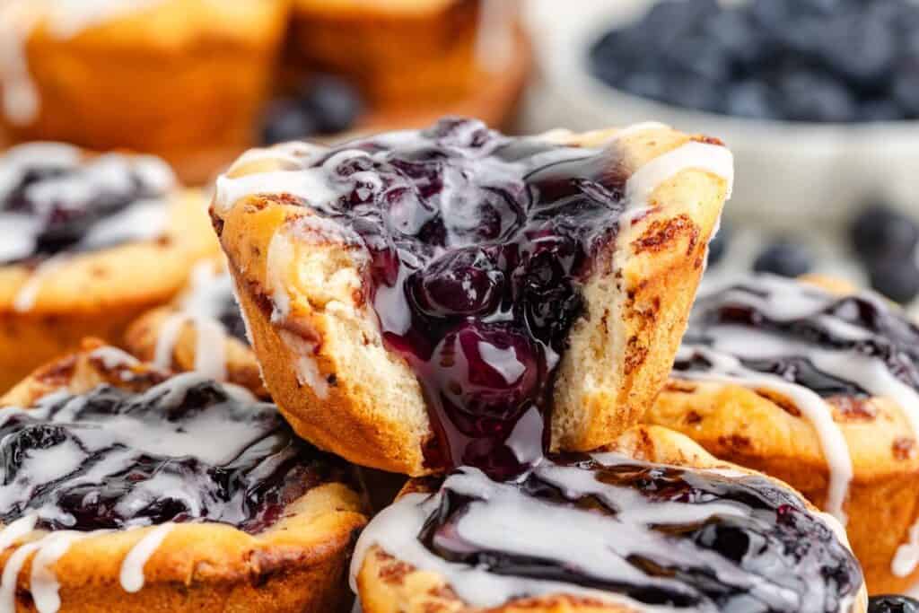 Stack of blueberry cinnamon rolls pies with icing; one pie is bit open, showing gooey blueberry filling inside.