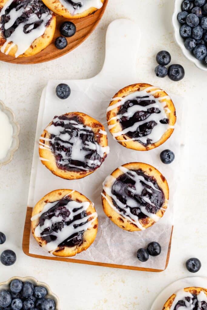 Four blueberry cinnamon roll pies with white icing on a white cutting board, surrounded by scattered fresh blueberries.