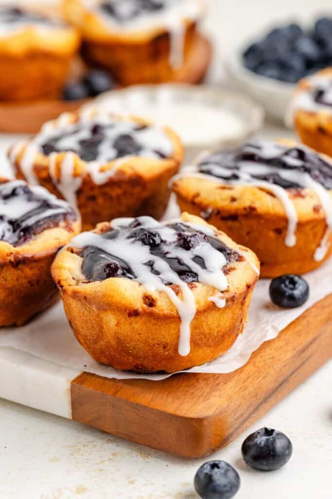 Blueberry mini pies with white icing on a wooden board, surrounded by fresh blueberries.