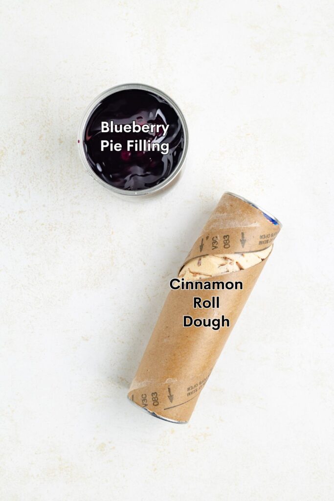 Overhead view of blueberry pie filling bowl beside a tube of cinnamon roll dough on light background.