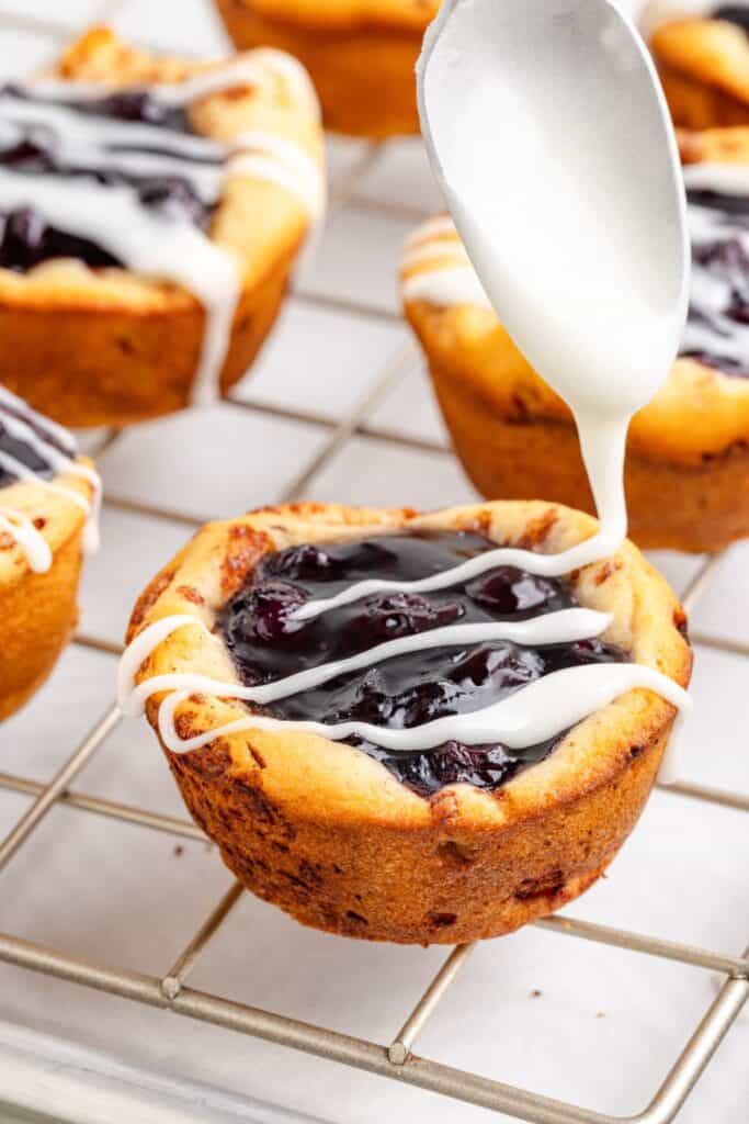 A spoon drizzles white icing onto a blueberry mini pie on a cooling rack.