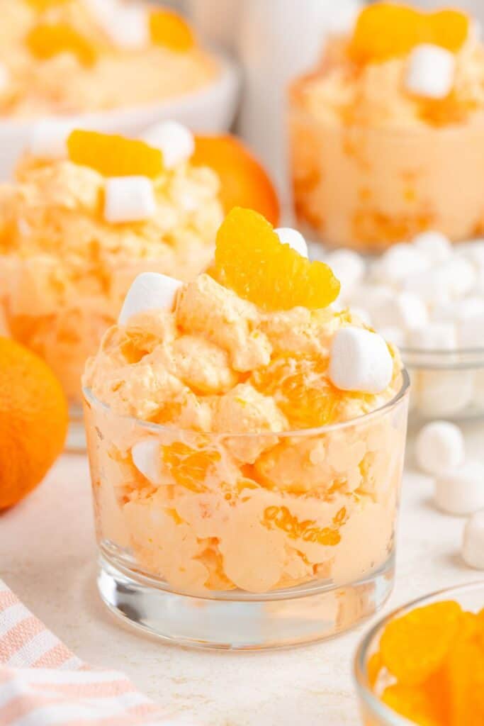 A glass dish of orange fluff salad topped with mandarin segments and mini marshmallows, with ingredients behind.