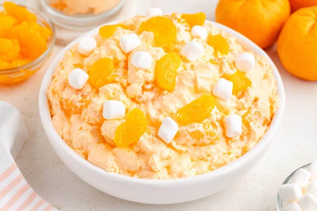 Bowl of orange fluff dessert with mandarin segments and marshmallows, tangerines and ingredients bowls in background.