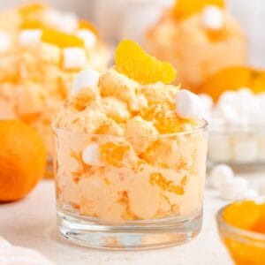 Glass bowl of orange fluff salad with mandarin slices and marshmallows; more bowls and mandarins in background.