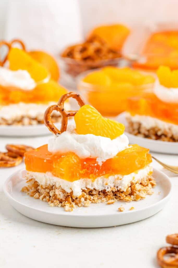 Layered orange pretzel salad with pretzel crust, creamy filling, orange gelatin, whipped topping, pretzel, and orange garnish.