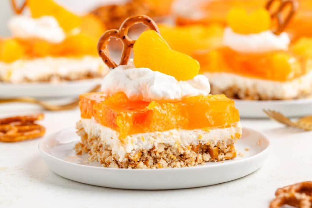 Mandarin Orange Pretzel Salad with pretzel crust, creamy filling, orange gelatin top, whipped cream, mandarin slice, and pretzel.