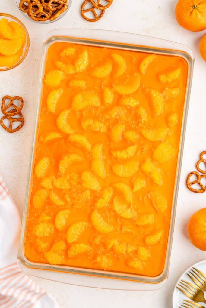 Glass dish of orange Jello salad with mandarin slices, surrounded by pretzels and fresh mandarin oranges.