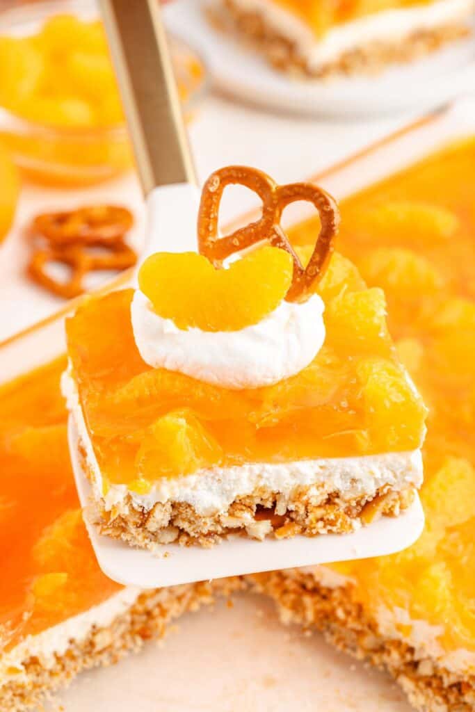 Square orange pretzel dessert slice with whipped cream, pretzel, and mandarin oranges, lifted on a spatula.