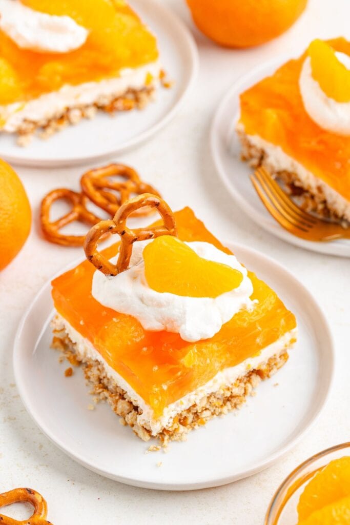 Slice of orange pretzel dessert salad with pretzel crust, whipped cream, and mandarin garnish on white plate; more slices nearby.