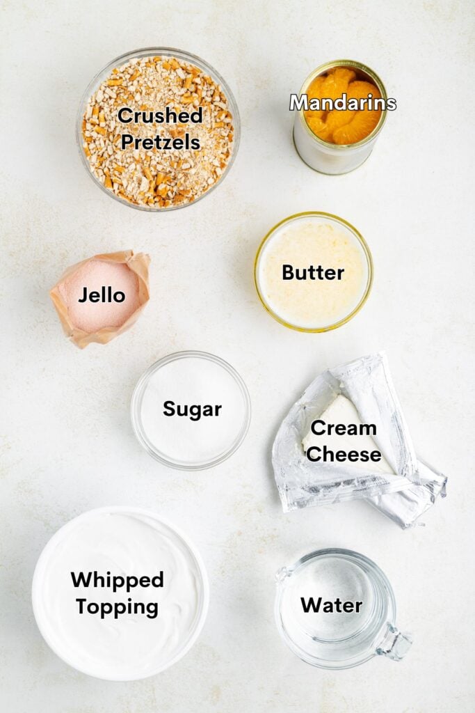Top-down view of bowls with crushed pretzels, mandarin oranges, jello, sugar, cream cheese, whipped topping, butter.