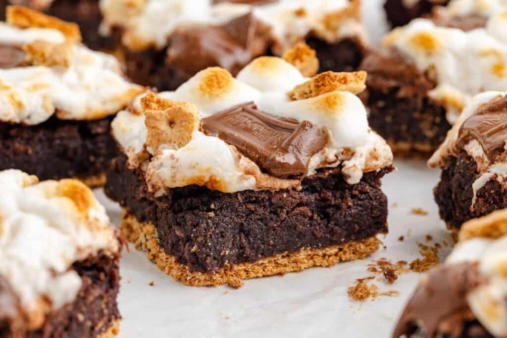 Close-up of a brownie topped with toasted marshmallows, chocolate pieces, and graham cracker crumbs, smores-style.