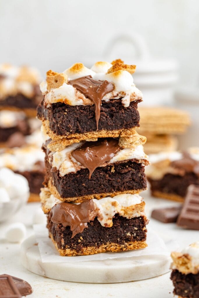 Stack of three smores brownies with melted chocolate and toasted marshmallows, chocolate pieces in background.