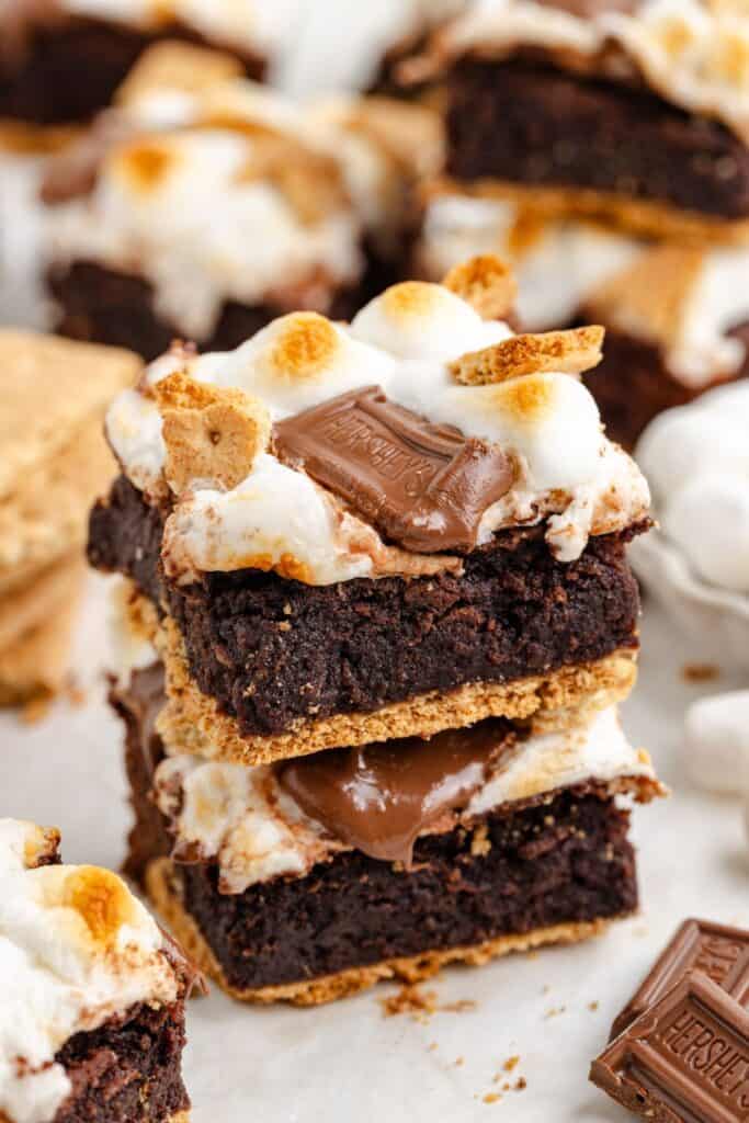 Stacked s'mores brownies topped with toasted marshmallows, chocolate, and graham crackers; more treats behind.