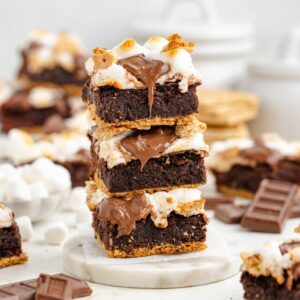 Stack of three s’mores brownies with melted chocolate and toasted marshmallows, brownies and chocolate in background.