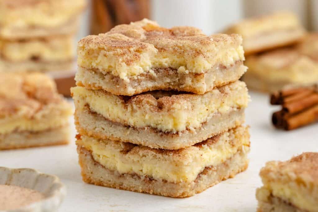 Stack of three layered Snickerdoodle dessert bars with creamy filling and cinnamon sugar topping; more bars and cinnamon sticks behind.