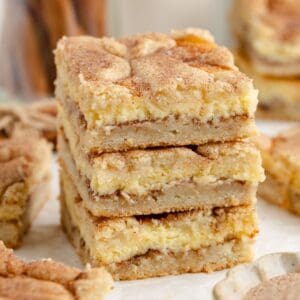 Three Snickerdoodle cheesecake bars with crumbly cinnamon sugar topping stacked on parchment, more bars blurred in background.