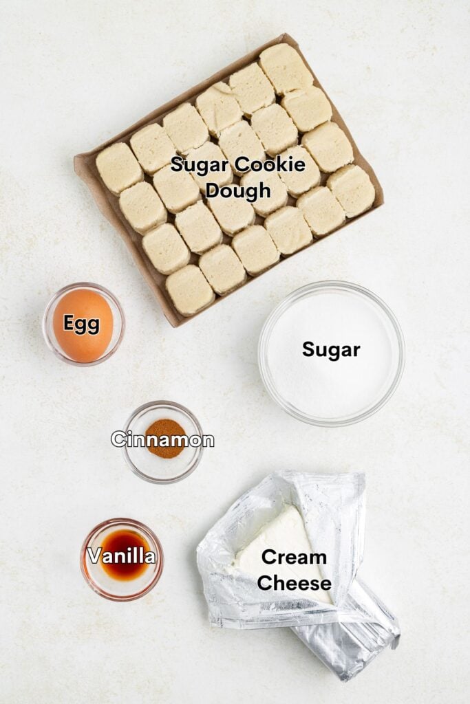 Tray of cubed sugar cookie dough with sugar, egg, cinnamon, vanilla, and opened cream cheese on light surface.