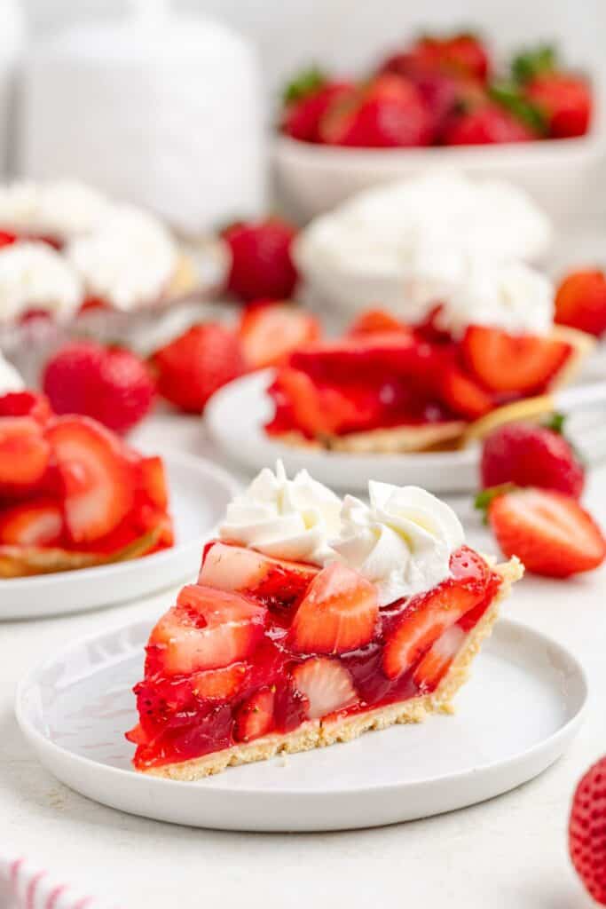 A slice of strawberry Jello pie with whipped cream on a white plate, with strawberries and more pie behind.