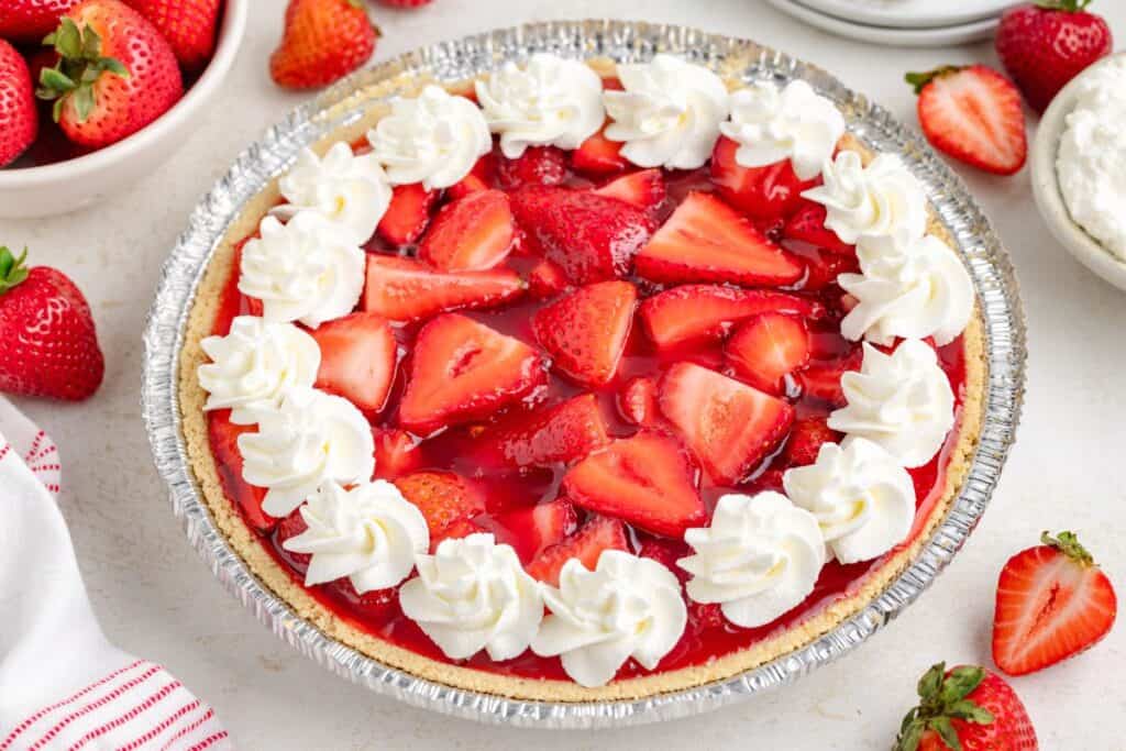 Jello strawberry pie with whipped cream swirls, surrounded by bowls of fresh strawberries and cream on the table.