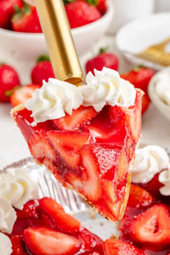 A slice of no-bake strawberry Jello pie with whipped cream and fresh strawberries is lifted from a tin.