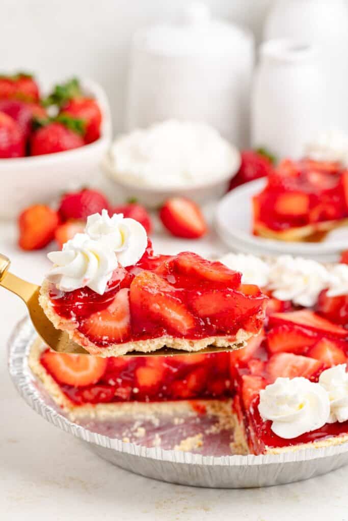 A slice of Jello strawberry pie with whipped cream is lifted from a tin; more slices and strawberries behind.
