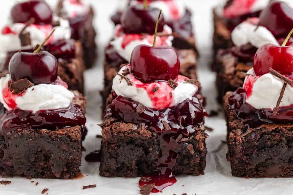 Chocolate cherry brownies topped with whipped cream, cherry sauce, chocolate shavings, and a whole cherry on each.