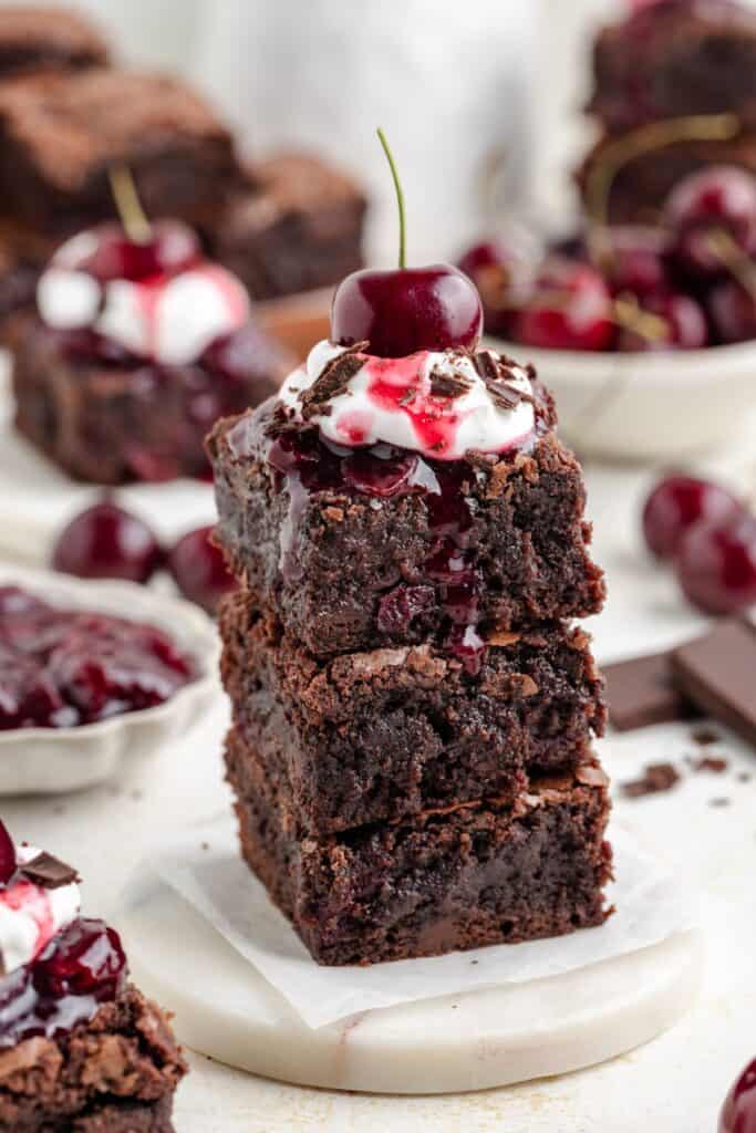 Stack of black forest brownies topped with whipped cream, cherry sauce, chocolate shavings, and a fresh cherry; more behind.