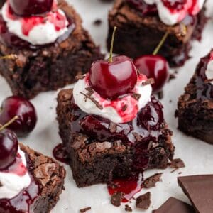 Black forest brownies with whipped cream, cherry sauce, chocolate shavings, and cherries on a white surface; chocolate pieces nearby.