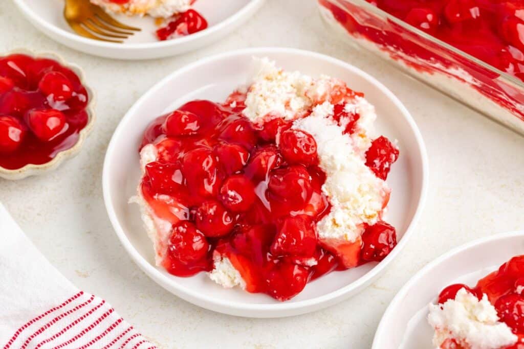 No Bake Cherry Dessert on white plate, covered in red sauce; other plates and baking dish in background.