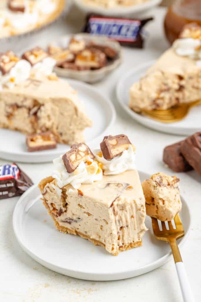 Slice of creamy Snickers pie with whipped cream and Snickers pieces on a plate, extra slices in background.