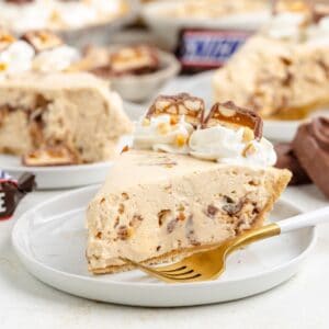 Slice of Snickers no bake pie with whipped cream, chocolate, caramel, and candy bar chunks on a white plate.