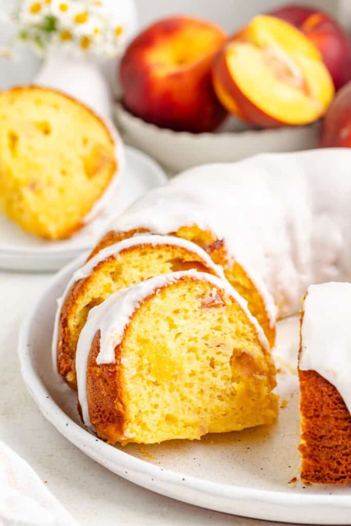 Sliced peach bundt cake with white icing on a plate, fresh peaches and flowers in the background.