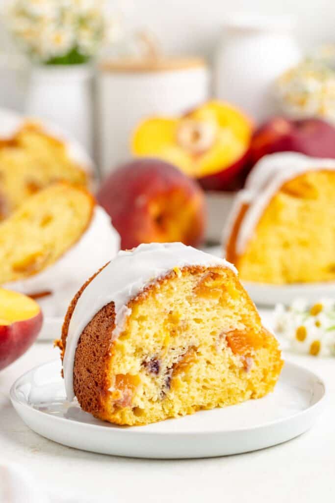A slice of peach bundt cake with white icing on a plate, cake and peaches in background.