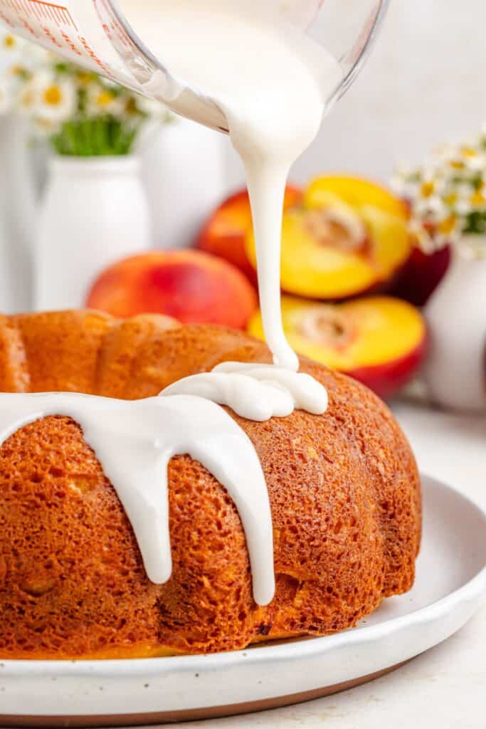 White glaze being poured onto a cake mix peach bundt cake; peaches and floral vases in the soft-focus background.