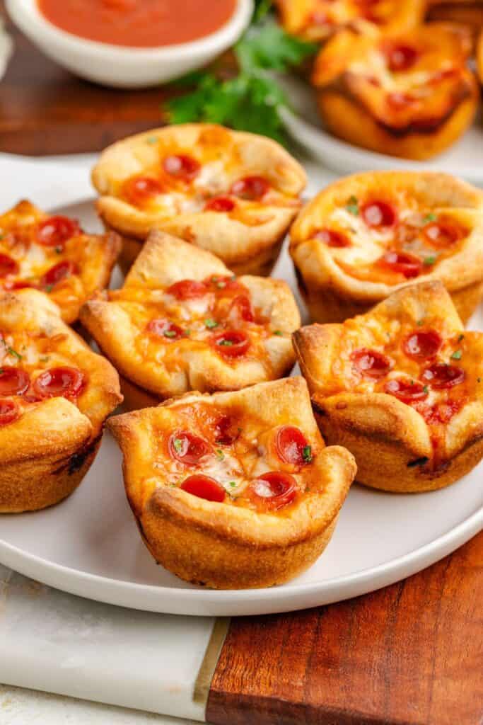 Mini pizza cups topped with melted cheese and pepperoni on a white plate, dipping sauce bowl behind.