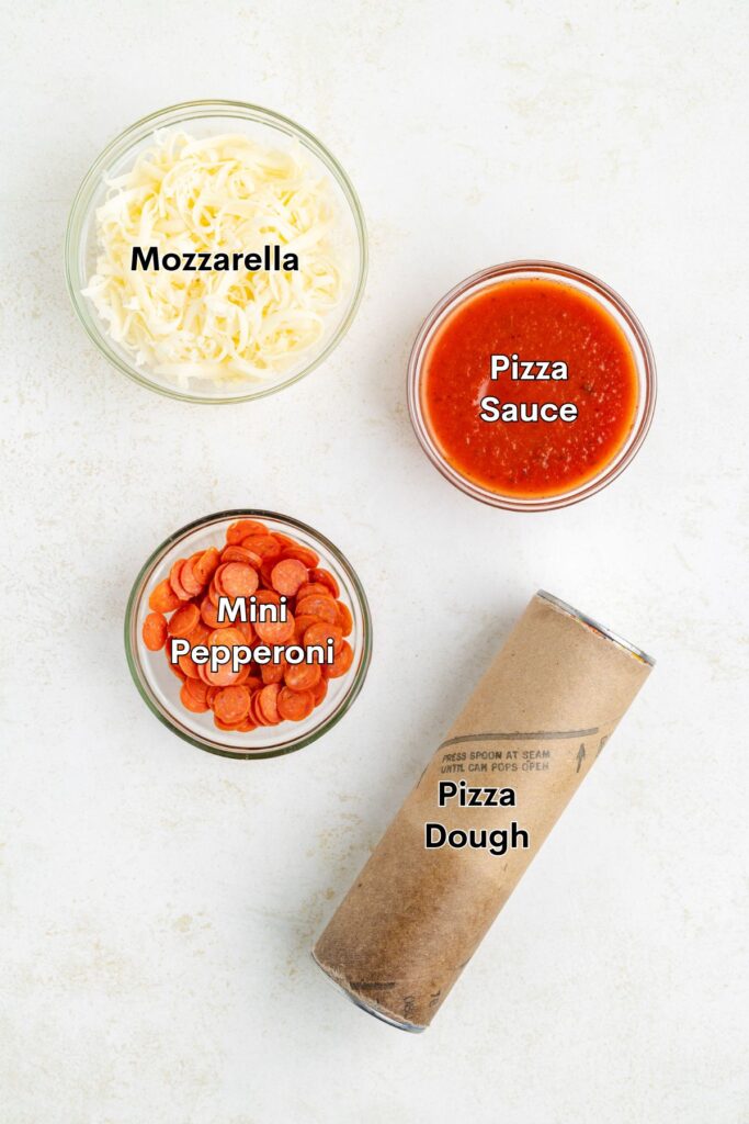 Four pizza ingredients: shredded mozzarella, sauce, mini pepperoni in bowls, and a tube of pizza dough on white surface.
