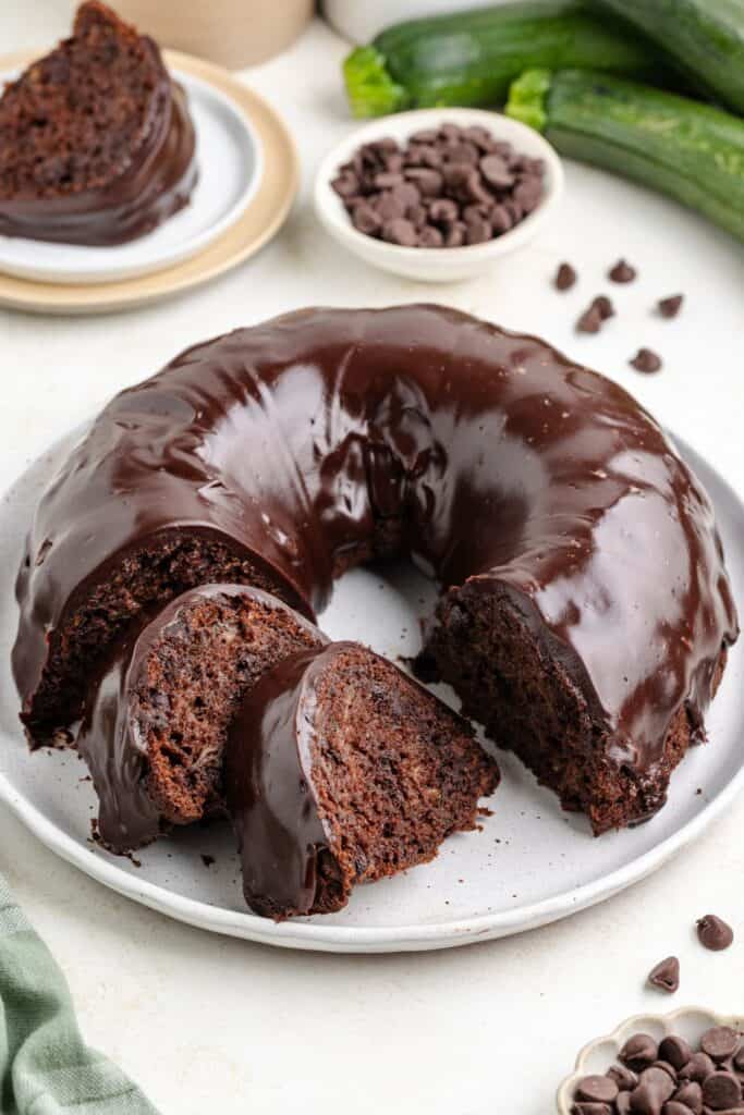 Sliced chocolate zucchini bundt cake with glaze on a plate, surrounded by chocolate chips and zucchinis.