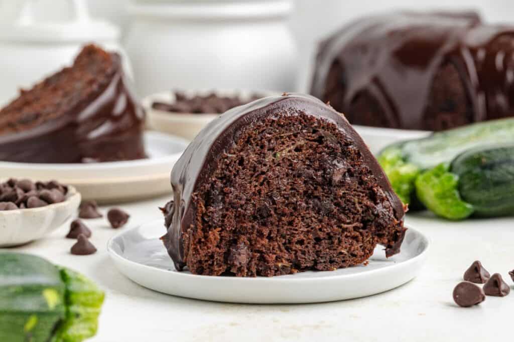 Slice of zucchini chocolate bundt cake with glossy glaze, on plate, surrounded by chocolate chips and fresh zucchini.