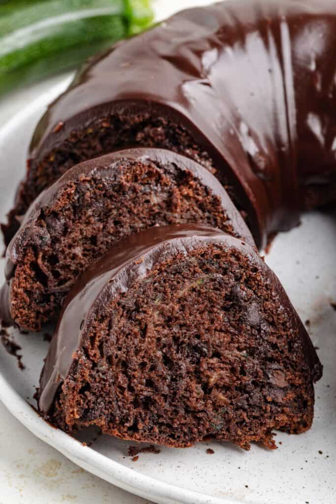 Chocolate bundt cake with glossy ganache, partially sliced to reveal moist, dark interior, on a white plate.