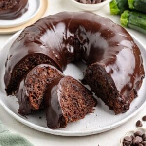 Sliced zucchini chocolate bundt cake with chocolate glaze on white plate; two slices placed in front of the cake.
