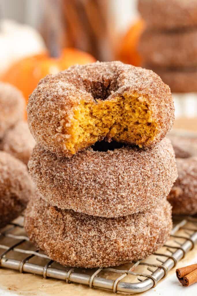 Three cinnamon sugar-pumpkin donuts stacked on a wire rack; top donut has a bite showing orange interior.