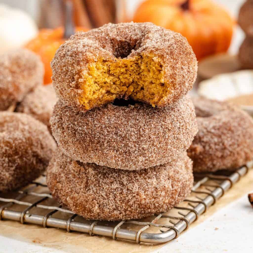 Stack of three cinnamon sugar pumpkin donuts on a cooling rack, top donut bitten, pumpkin blurred in background.